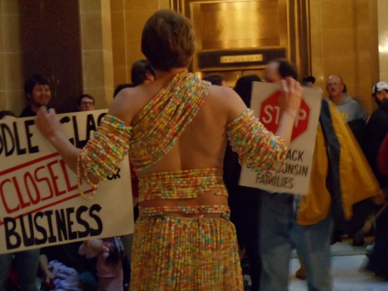 The front of this UW-Madison student's sign says "Even I Look Less Ridiculous Than Scott Walker." He is wearing a "grass skirt" and toga-type top made entirely of candy necklaces. The back of the sign: Middle Class: CLOSED FOR BUSINESS