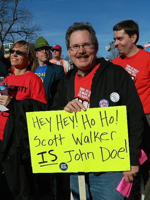 man holding a sign saying "hey hey! ho ho! Scott Walker is John Doe!"